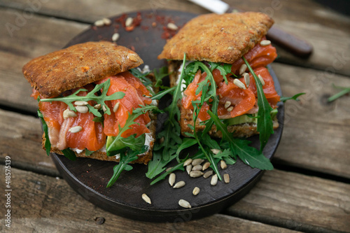 A delicious snack for lunch. Delicious sandwich with lightly salted salmon. With a lush bun, with arugula greens and sunflower seeds! Served on a wooden platter and rustic background. Horizontal orien