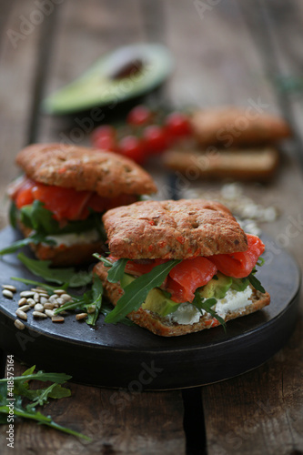 A delicious snack for lunch. Delicious sandwich with lightly salted salmon. With a lush bun, with arugula greens and sunflower seeds! Served on a wooden platter and rustic background. Horizontal orien