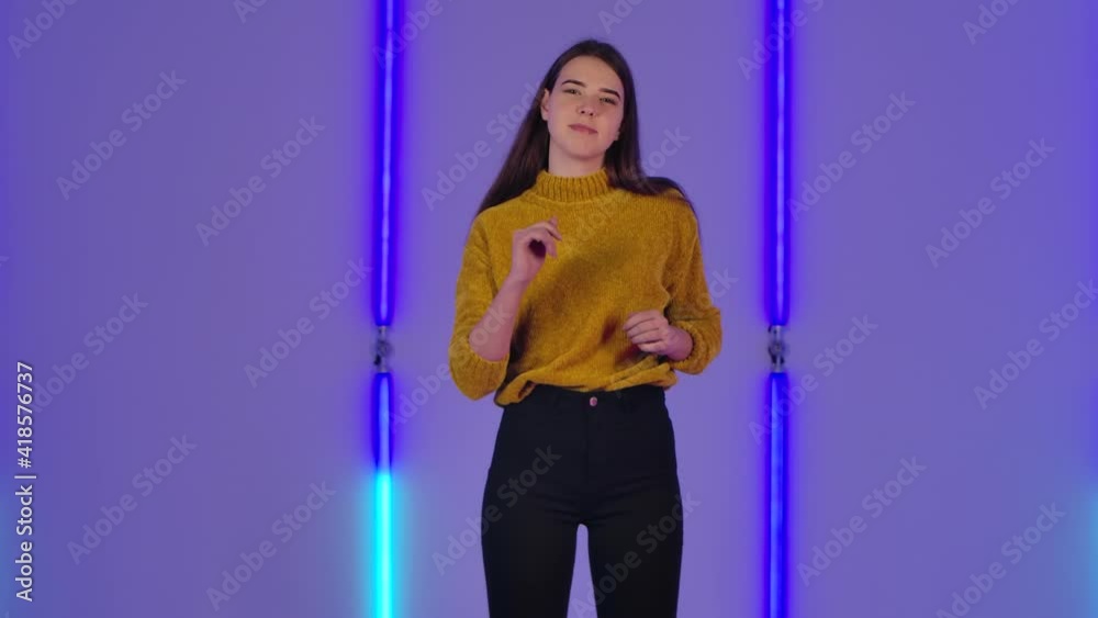Portrait of young cute woman poses on camera. Fashion model with long hair in mustard sweater against background of bright neon lights in dark studio. Slow motion. Portrait of young cute woman poses on camera. Fashion model with long hair in mustard sweater against background of bright neon lights in dark studio. Slow motion.