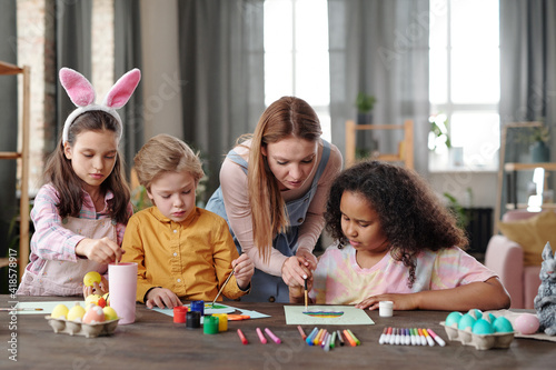 Pretty young blond woman helping cute kids with painting picture of Easter egg