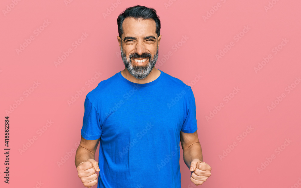 Middle aged man with beard wearing casual blue t shirt very happy and excited doing winner gesture with arms raised, smiling and screaming for success. celebration concept. Middle aged man with beard wearing casual blue t shirt very happy and excited doing winner gesture with arms raised, smiling and screaming for success. celebration concept.