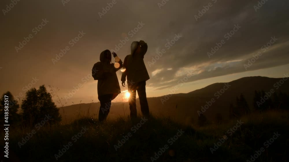 road Two hikers in yellow jackets climb to the top of the mountain holding hands in the rain. Woman helps a friend to raise his hand to the top against the backdrop of a mountain landscape at sunset.