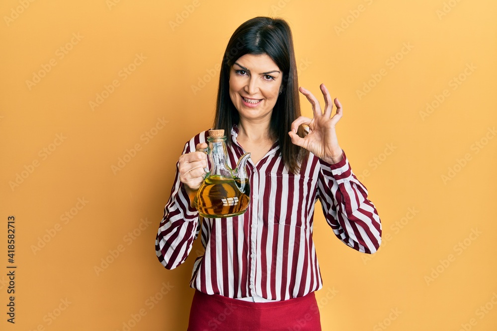 Young hispanic woman holding olive oil can doing ok sign with fingers, smiling friendly gesturing excellent symbol Young hispanic woman holding olive oil can doing ok sign with fingers, smiling friendly gesturing excellent symbol