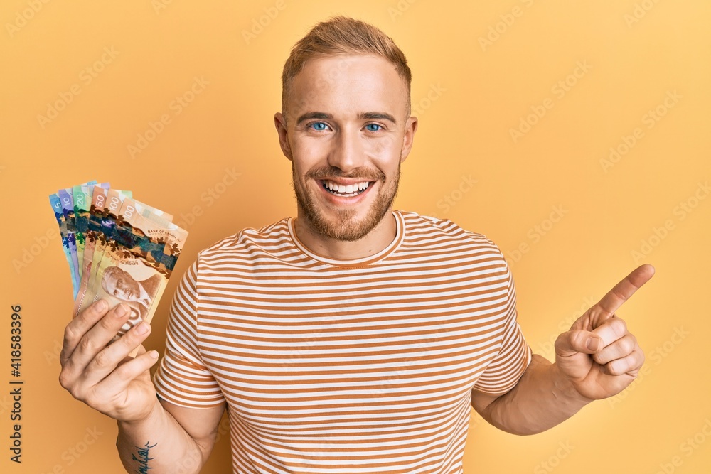Young caucasian man holding canadian dollars smiling happy pointing with hand and finger to the side