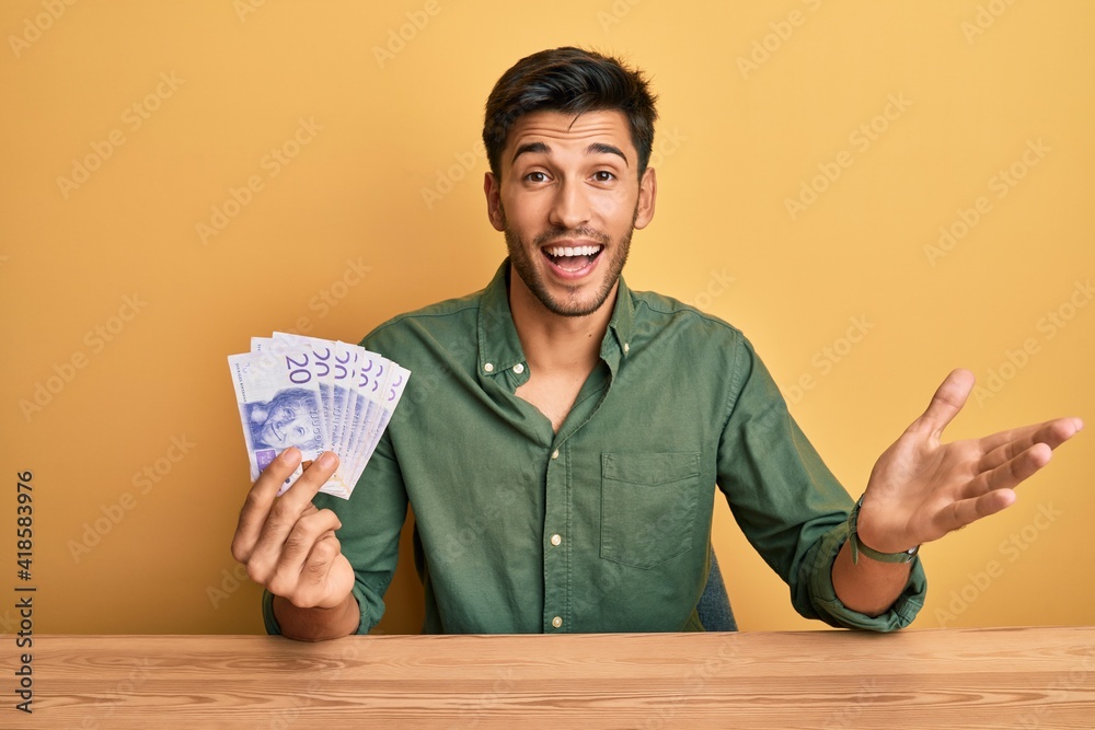 Young handsome man holding 20 swedish krona banknotes celebrating achievement with happy smile and winner expression with raised hand Young handsome man holding 20 swedish krona banknotes celebrating achievement with happy smile and winner expression with raised hand