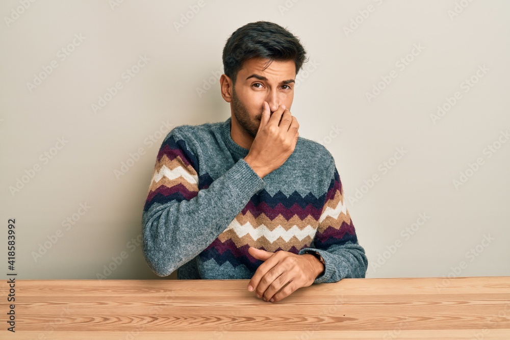 Young handsome man wearing casual sweater sitting on the table smelling something stinky and disgusting, intolerable smell, holding breath with fingers on nose. bad smell Young handsome man wearing casual sweater sitting on the table smelling something stinky and disgusting, intolerable smell, holding breath with fingers on nose. bad smell