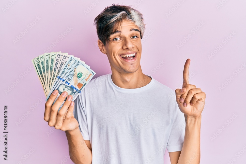Young hispanic man holding dollars smiling with an idea or question pointing finger with happy face, number one Young hispanic man holding dollars smiling with an idea or question pointing finger with happy face, number one