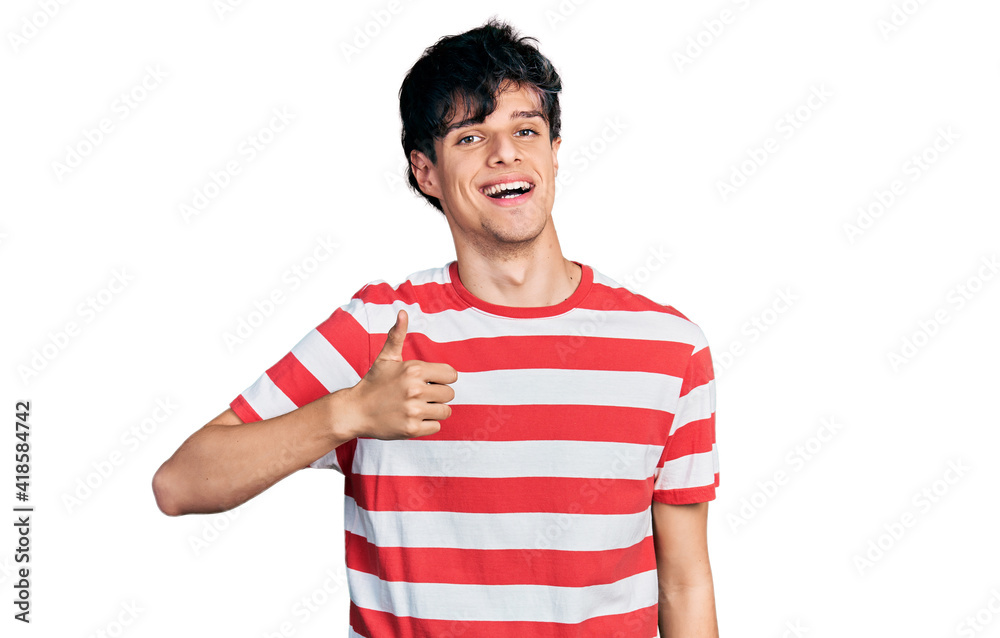 Handsome hipster young man wearing casual striped t shirt doing happy thumbs up gesture with hand. approving expression looking at the camera showing success.