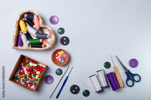 Wooden heart shaped box of multicolored beads and buttons, awl, scissors, crochets and spool of threads for sewing and embroidery. Set of materials for handcraft, making of bijouterie and accessories