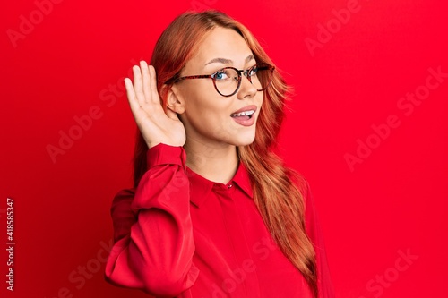 Young beautiful redhead woman wearing casual clothes and glasses over red background smiling with hand over ear listening an hearing to rumor or gossip. deafness concept.