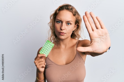 Beautiful caucasian woman holding birth control pills with open hand doing stop sign with serious and confident expression, defense gesture
