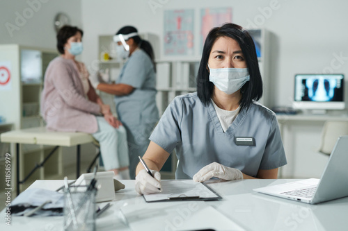 Young brunette female clinician in protective workwear sitting by desk in front of laptop