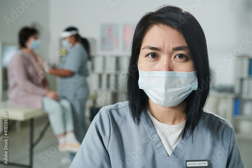 Young serious Asian female clinician in uniform and protective mask looking at you