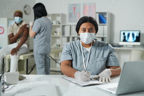 Young mixed-race clinician in protective workwear filling in medical document by desk