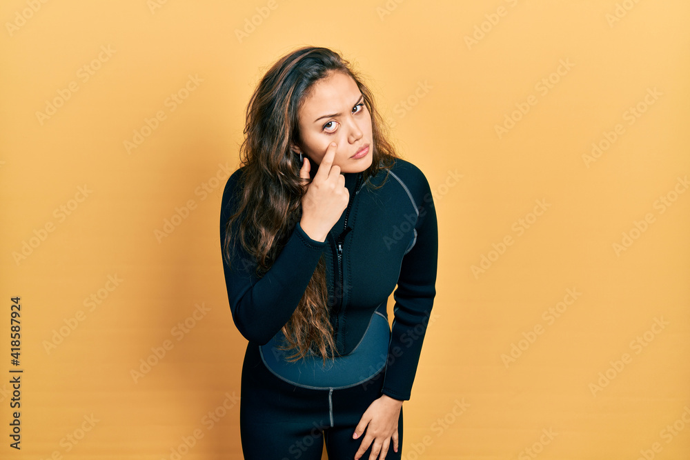 Young hispanic girl wearing diver neoprene uniform pointing to the eye watching you gesture, suspicious expression Young hispanic girl wearing diver neoprene uniform pointing to the eye watching you gesture, suspicious expression