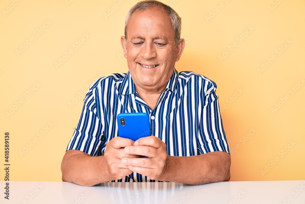 Senior handsome man with gray hair using smartphone sitting on the table looking positive and happy standing and smiling with a confident smile showing teeth Senior handsome man with gray hair using smartphone sitting on the table looking positive and happy standing and smiling with a confident smile showing teeth