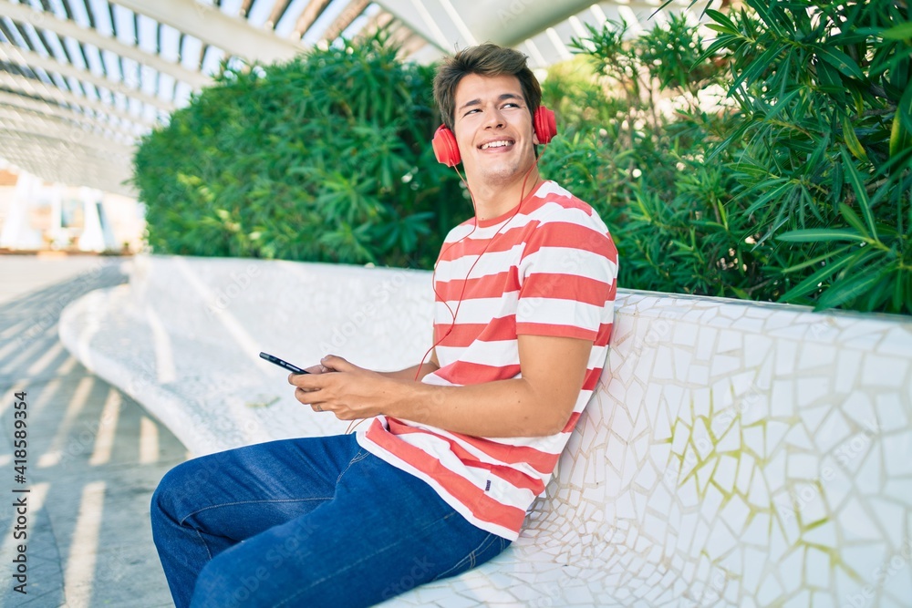 Fototapeta premium Young caucasian man smiling happy using smartphone and headphones sitting at the park.