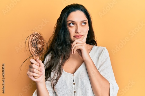 Beautiful middle eastern woman holding comb loosing hair serious face thinking about question with hand on chin, thoughtful about confusing idea photo