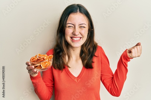 Young brunette woman holding bowl of dry orange screaming proud, celebrating victory and success very excited with raised arm