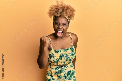 Young african woman with afro hair wearing summer dress angry and mad raising fist frustrated and furious while shouting with anger. rage and aggressive concept.