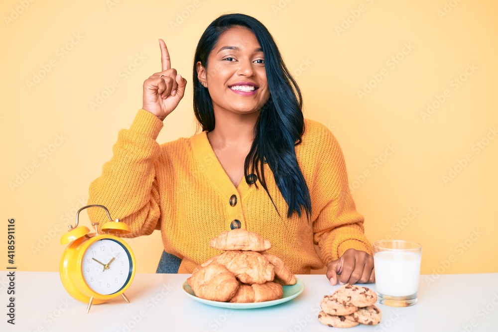 Beautiful latin young woman with long hair sitting on the table having breakfast pointing finger up with successful idea. exited and happy. number one. Beautiful latin young woman with long hair sitting on the table having breakfast pointing finger up with successful idea. exited and happy. number one.