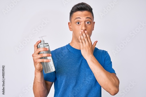 Young hispanic boy using hand sanitizer gel covering mouth with hand, shocked and afraid for mistake. surprised expression