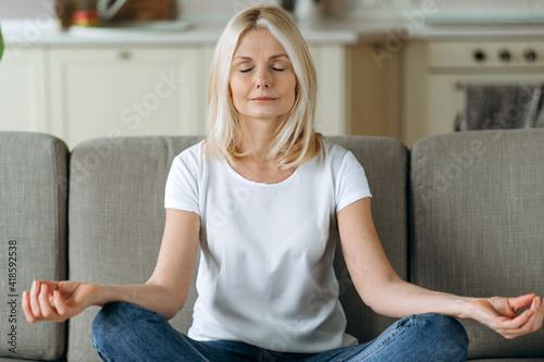 Calm attractive middle aged woman sits on sofa at home and chilling. Senior female is practise yoga and meditation in lotus position with eyes closed, relieve stress, calmness concept