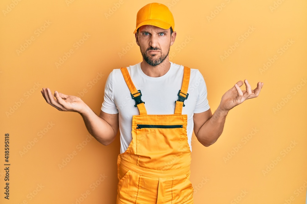 Young handsome man wearing handyman uniform over yellow background clueless and confused with open arms, no idea concept.