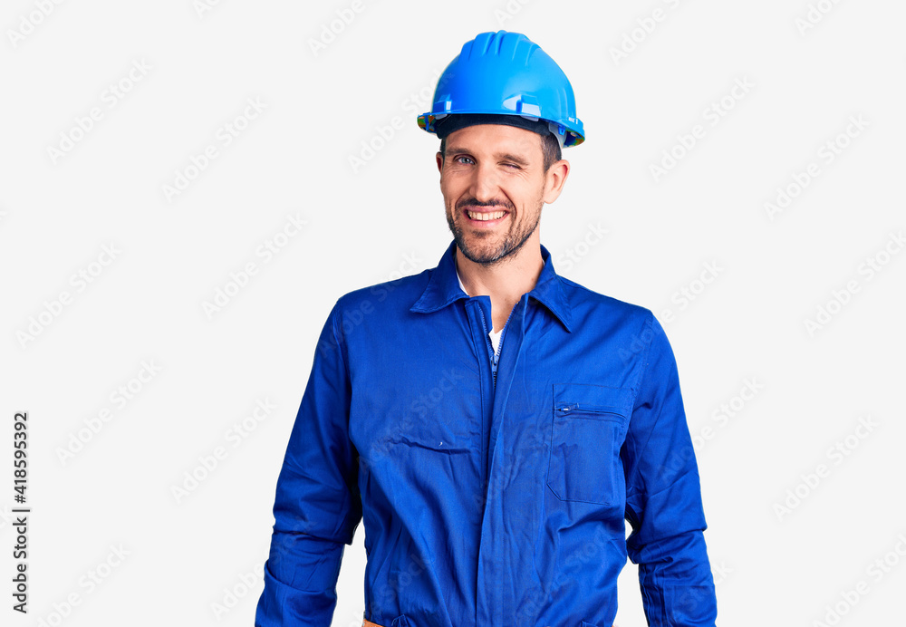 Young handsome man wearing worker uniform and hardhat winking looking at the camera with sexy expression, cheerful and happy face.