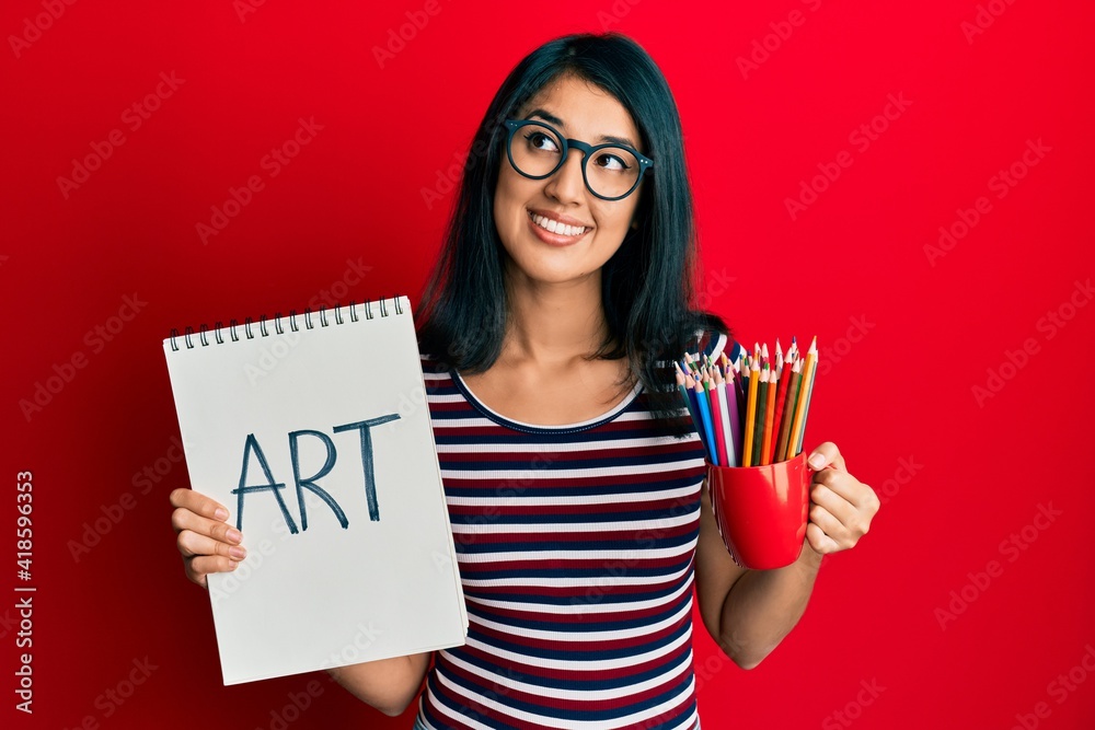 Beautiful asian young woman holding art notebook and colored pencils smiling looking to the side and staring away thinking. Beautiful asian young woman holding art notebook and colored pencils smiling looking to the side and staring away thinking.