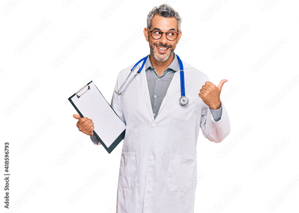 Middle age grey-haired man wearing doctor stethoscope holding clipboard pointing thumb up to the side smiling happy with open mouth