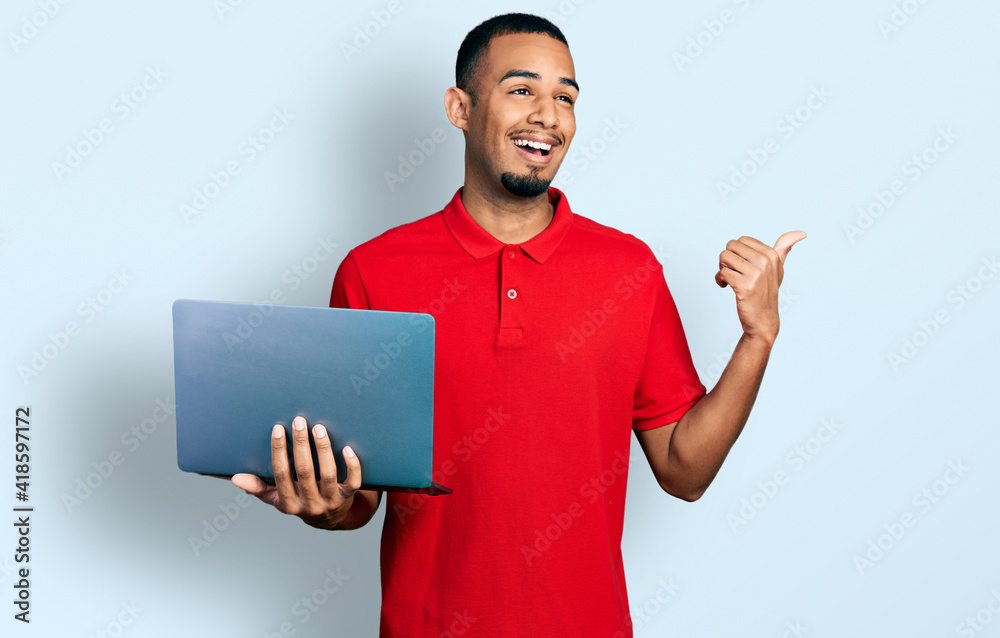 Young african american man working using computer laptop pointing thumb up to the side smiling happy with open mouth Young african american man working using computer laptop pointing thumb up to the side smiling happy with open mouth