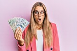 Young blonde woman wearing business style holding dollars scared and amazed with open mouth for surprise, disbelief face