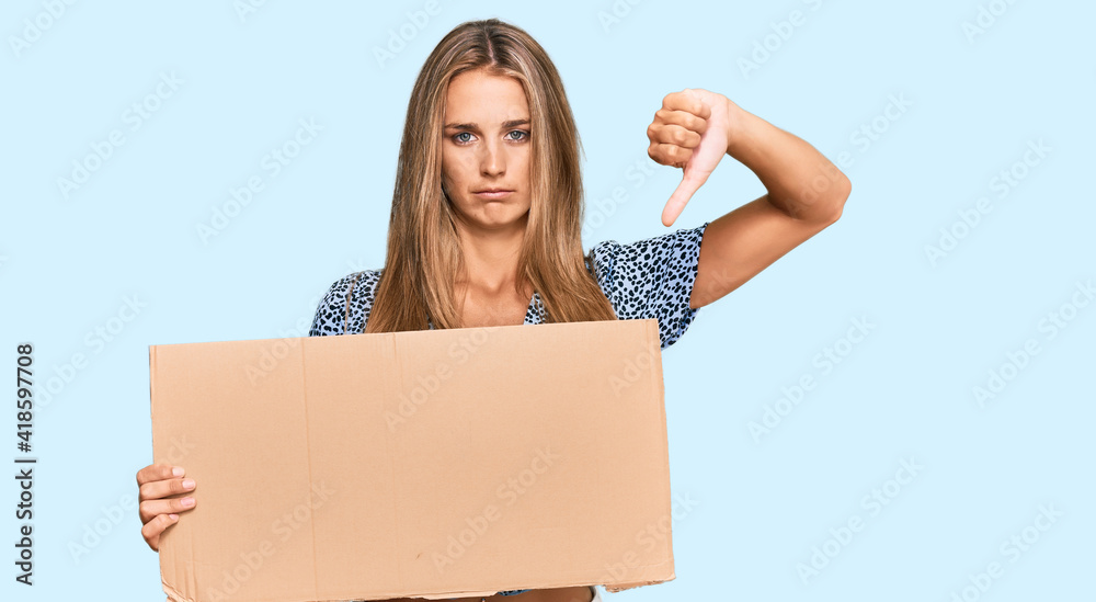 Young blonde woman holding cardboard empty banner with angry face, negative sign showing dislike with thumbs down, rejection concept Young blonde woman holding cardboard empty banner with angry face, negative sign showing dislike with thumbs down, rejection concept