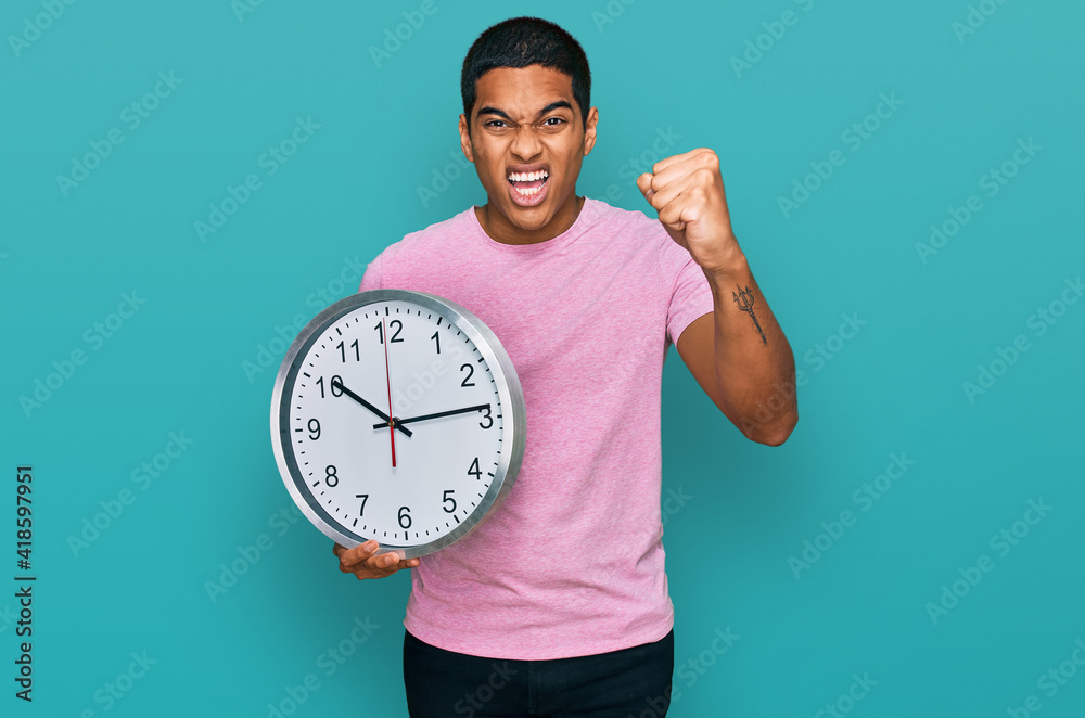 Young handsome hispanic man holding big clock annoyed and frustrated shouting with anger, yelling crazy with anger and hand raised Young handsome hispanic man holding big clock annoyed and frustrated shouting with anger, yelling crazy with anger and hand raised