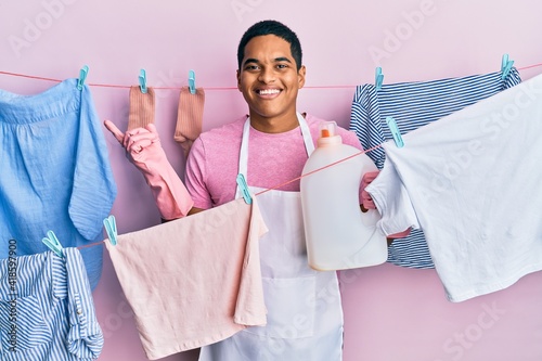 Young handsome hispanic man doing laundry holding detergent bottle smiling happy pointing with hand and finger to the side