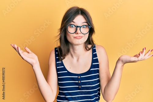 Foto Young beautiful caucasian girl wearing casual clothes and glasses clueless and confused with open arms, no idea and doubtful face
