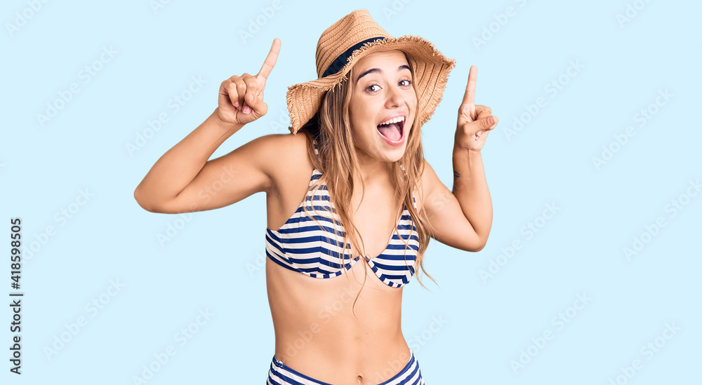 Young beautiful blonde woman wearing bikini and hat smiling amazed and surprised and pointing up with fingers and raised arms. Young beautiful blonde woman wearing bikini and hat smiling amazed and surprised and pointing up with fingers and raised arms.