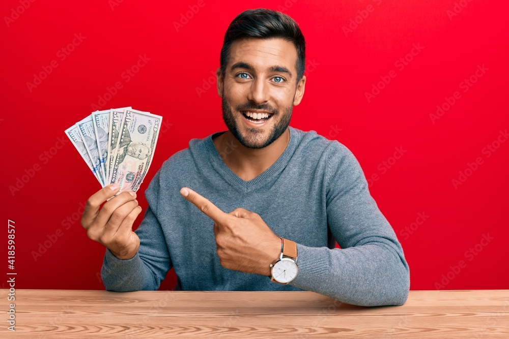 Handsome hispanic man holding dollars smiling happy pointing with hand and finger Handsome hispanic man holding dollars smiling happy pointing with hand and finger