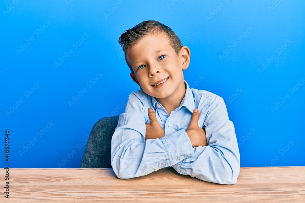 Adorable caucasian kid wearing casual clothes sitting on the table happy face smiling with crossed arms looking at the camera. positive person. Adorable caucasian kid wearing casual clothes sitting on the table happy face smiling with crossed arms looking at the camera. positive person.