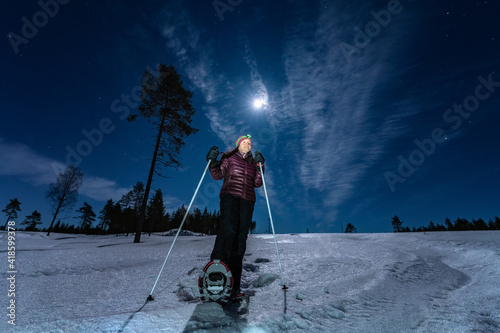 Beautiful middle aged Caucasian women in magenta haze jacket and snow shoes stands in night rare snowy winter forest under full moon light and looks to left side up. Night walk, Lapland, Umea, Sweden