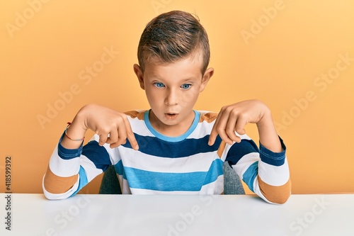 Adorable caucasian kid wearing casual clothes sitting on the table pointing down with fingers showing advertisement, surprised face and open mouth