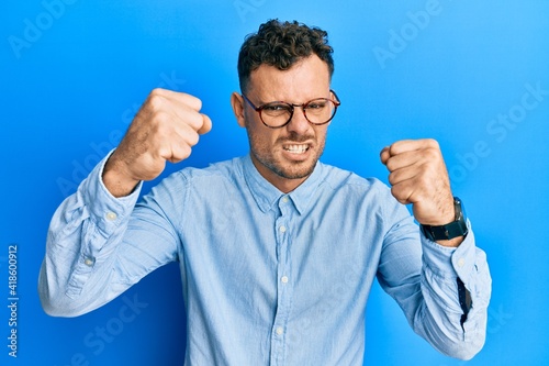 Young hispanic man wearing casual clothes and glasses angry and mad raising fists frustrated and furious while shouting with anger. rage and aggressive concept.