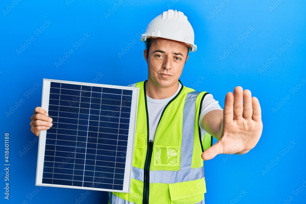 Handsome young man holding photovoltaic solar panel with open hand doing stop sign with serious and confident expression, defense gesture Handsome young man holding photovoltaic solar panel with open hand doing stop sign with serious and confident expression, defense gesture