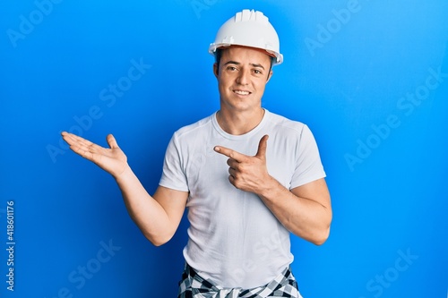 Handsome young man wearing builder uniform and hardhat amazed and smiling to the camera while presenting with hand and pointing with finger.