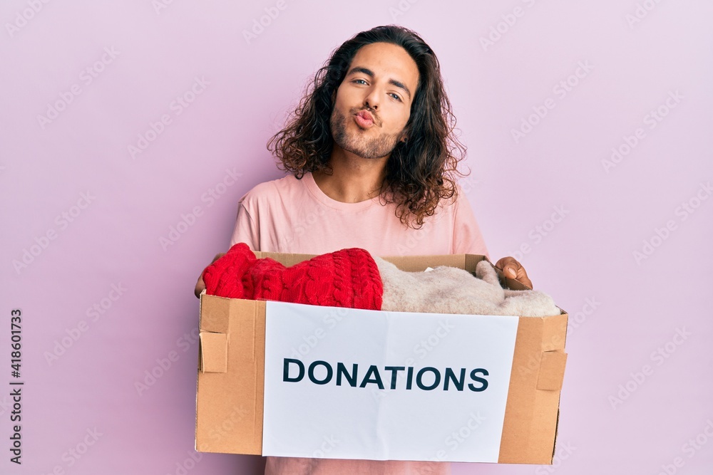 Young handsome man with long hair holding donations box for charity looking at the camera blowing a kiss being lovely and sexy. love expression. Young handsome man with long hair holding donations box for charity looking at the camera blowing a kiss being lovely and sexy. love expression.