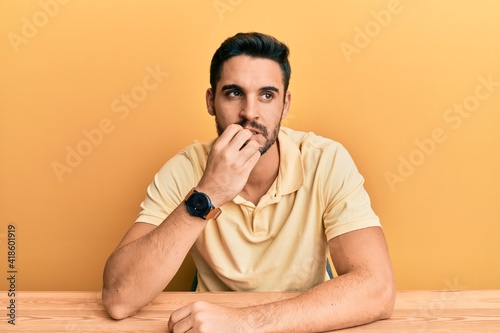 Young hispanic man wearing casual clothes sitting on the table looking stressed and nervous with hands on mouth biting nails. anxiety problem.