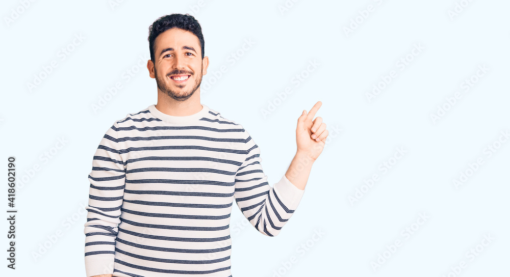 Young hispanic man wearing casual clothes with a big smile on face, pointing with hand finger to the side looking at the camera. Young hispanic man wearing casual clothes with a big smile on face, pointing with hand finger to the side looking at the camera.