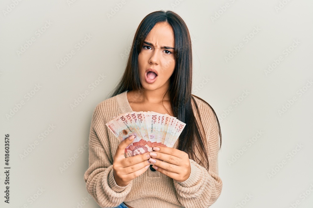Young beautiful hispanic girl holding 10 colombian pesos banknotes in shock face, looking skeptical and sarcastic, surprised with open mouth Young beautiful hispanic girl holding 10 colombian pesos banknotes in shock face, looking skeptical and sarcastic, surprised with open mouth