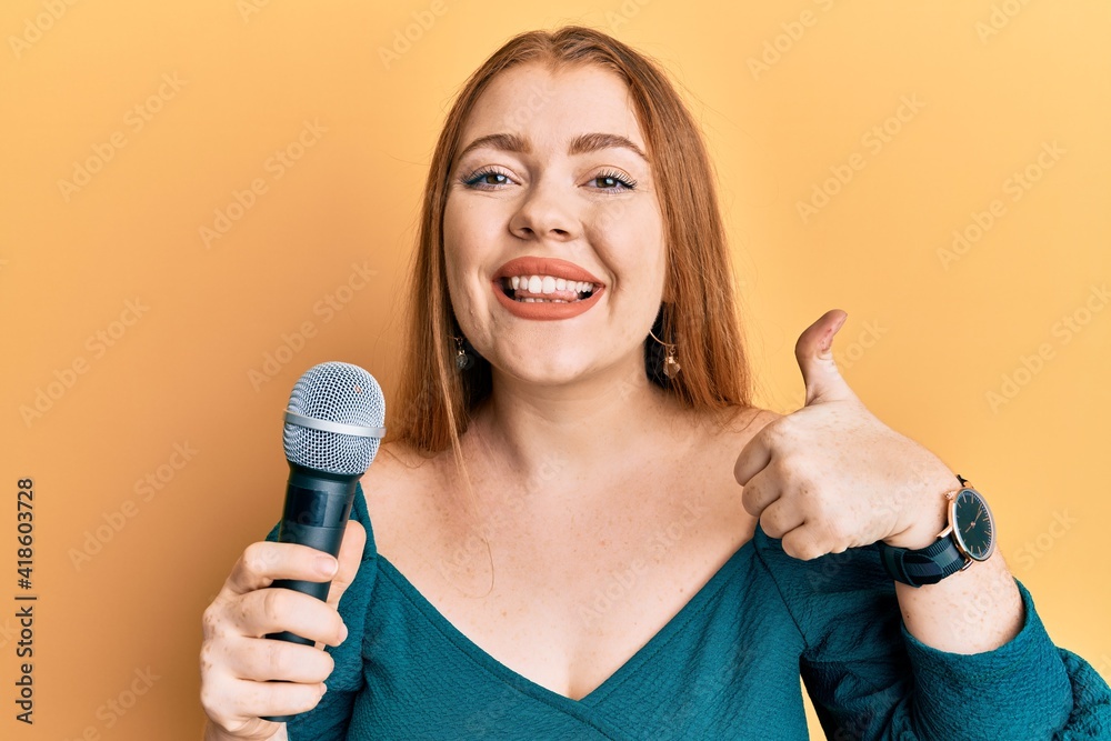 Young beautiful redhead woman singing song using microphone smiling happy and positive, thumb up doing excellent and approval sign Young beautiful redhead woman singing song using microphone smiling happy and positive, thumb up doing excellent and approval sign